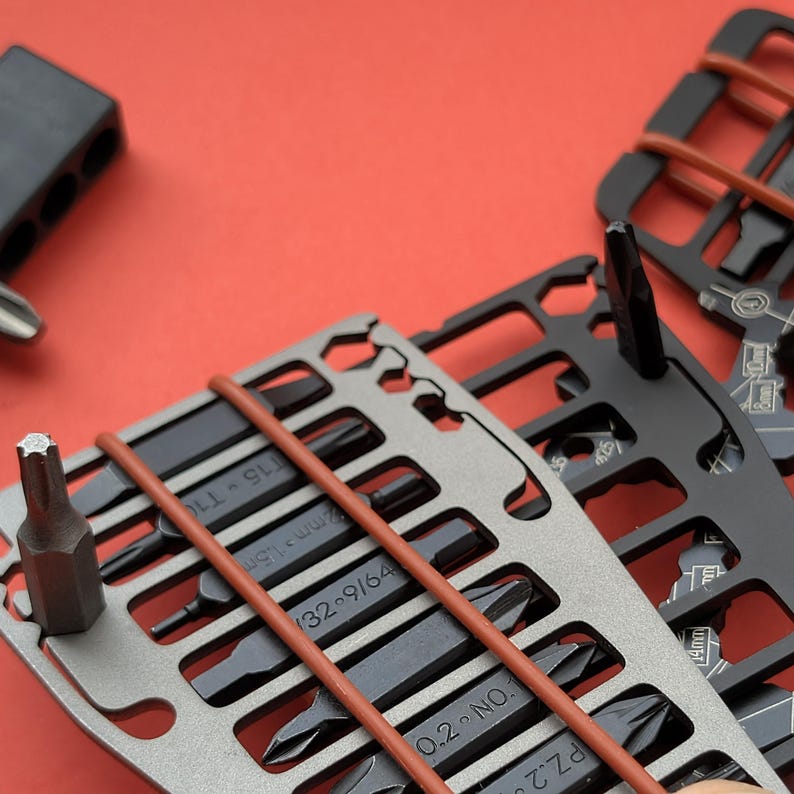 May include: Close-up of a set of multi-tool cards with various screwdriver bits. The cards are silver and black, held together with brown rubber bands, and set against a red background. The bits are black and arranged in rows.