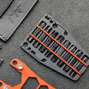 May include: A close-up shot of a black multi-tool bit set with a red rubber band holding the bits in place. The bits are various sizes and shapes. An orange tool and a black leather case are also visible.