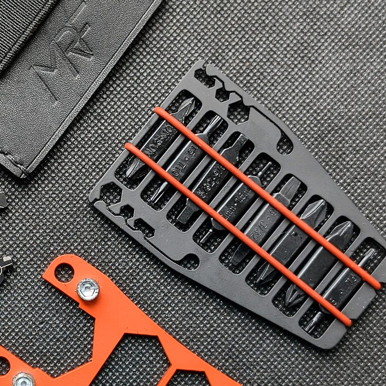 May include: A close-up shot of a black multi-tool bit set with a red rubber band holding the bits in place. The bits are various sizes and shapes. An orange tool and a black leather case are also visible.