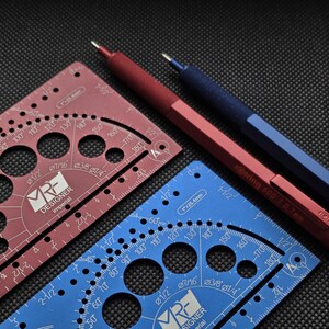 May include: A red and a blue metal drafting template, each featuring circular cutouts, degree markings, and the text 'MR DESIGNER imperial'. Beside them lie a red and a blue mechanical pencil, with the red one labeled 'rotring 600 | 0.7mm'.