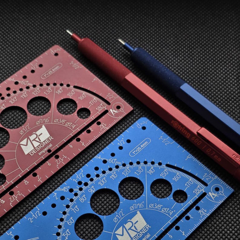 May include: A red and a blue metal drafting template, each featuring circular cutouts, degree markings, and the text 'MR DESIGNER imperial'. Beside them lie a red and a blue mechanical pencil, with the red one labeled 'rotring 600 | 0.7mm'.