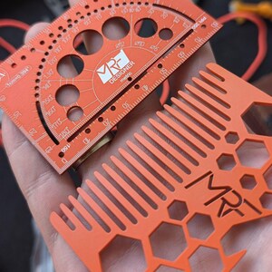 May include: An orange metric designer tool featuring a protractor, circular cutouts, and a ruler, alongside an orange comb with a hexagonal pattern and the MRF logo. Both items are made of a flat, matte material with white printed markings.