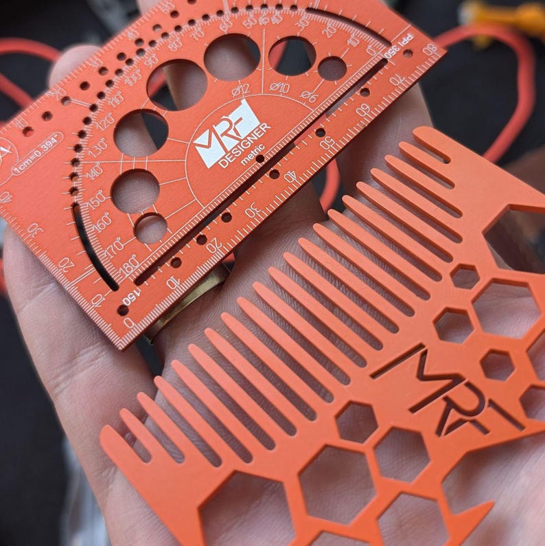 May include: An orange metric designer tool featuring a protractor, circular cutouts, and a ruler, alongside an orange comb with a hexagonal pattern and the MRF logo. Both items are made of a flat, matte material with white printed markings.
