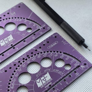 May include: Two purple metal drafting templates with white markings, including circular cutouts, degree scales, and imperial measurements labeled MRF DESIGNER. A black mechanical pencil with a textured grip rests diagonally across the top.
