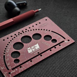 May include: A maroon metal drafting tool featuring a protractor, circular cutouts, and imperial measurement markings. The tool is labeled with the text MRF DESIGNER imperial and 1 inch equals 25.4mm, positioned near a maroon pen and black earbuds.