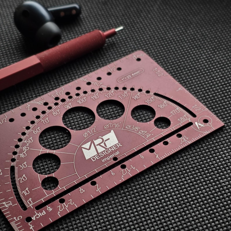 May include: A maroon metal drafting tool featuring a protractor, circular cutouts, and imperial measurement markings. The tool is labeled with the text MRF DESIGNER imperial and 1 inch equals 25.4mm, positioned near a maroon pen and black earbuds.