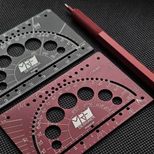 May include: Two metal drafting templates and a red mechanical pencil. One template is black with metric markings, and the other is maroon with imperial markings. Both feature circular cutouts, protractor scales, and the MRF Designer logo.