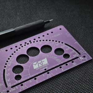 May include: A purple metal drafting template with a protractor, circular cutouts, and imperial measurement markings. The tool features the text MRF DESIGNER imperial and 1 inch equals 25.4 mm. A black mechanical pencil rests above the tool.