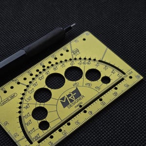 May include: A gold-colored metal drafting tool featuring a protractor, various circular cutouts labeled with diameters, and a ruler edge. The tool is marked with the text MRF DESIGNER metric, 1cm=0.394, and PPI 350. A black mechanical pencil rests nearby.