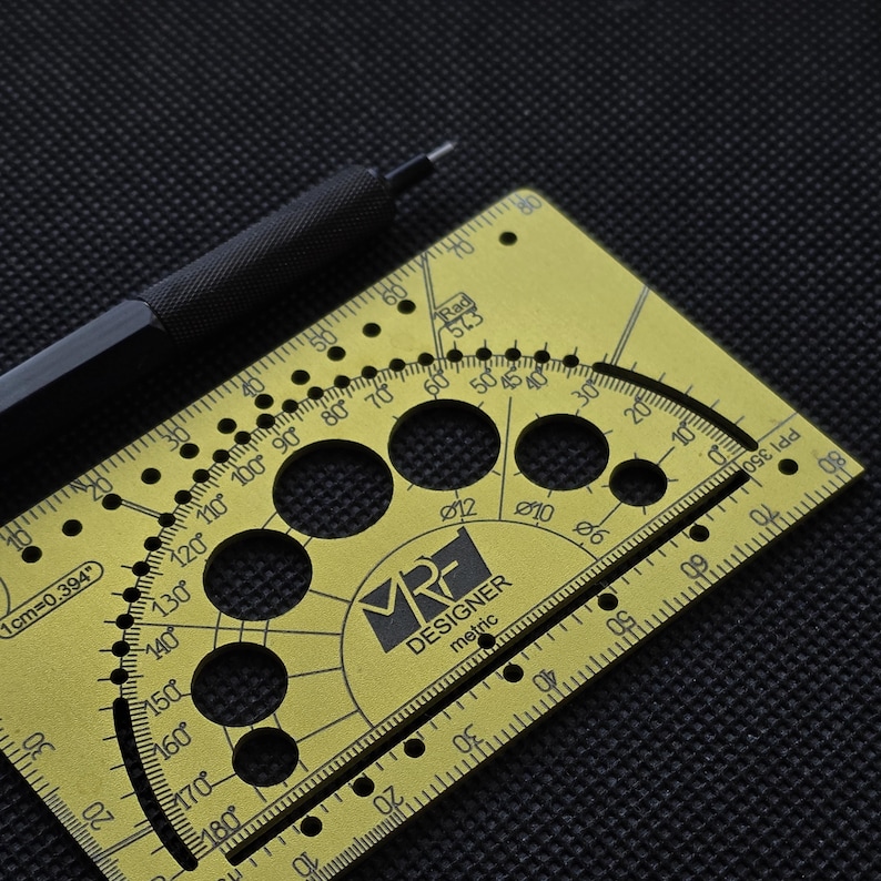 May include: A gold-colored metal drafting tool featuring a protractor, various circular cutouts labeled with diameters, and a ruler edge. The tool is marked with the text MRF DESIGNER metric, 1cm=0.394, and PPI 350. A black mechanical pencil rests nearby.