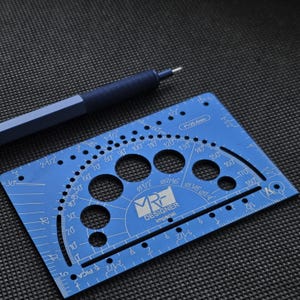 May include: A blue metal drafting tool featuring a protractor, circular cutouts, and imperial measurement markings, accompanied by a matching blue mechanical pencil with a textured grip. The tool is labeled MRF DESIGNER imperial.