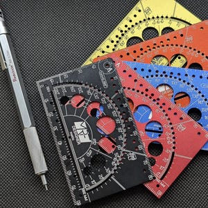 May include: A silver Redcircle 600 0.7mm mechanical pencil positioned next to a stack of four rectangular metal drafting templates in black, red, blue, and yellow. Each template features circular cutouts, degree markings, and the text MR DESIGNER.
