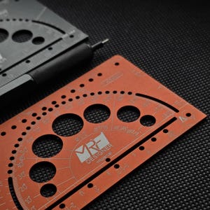 May include: A copper-colored metal drafting template featuring circular cutouts, angle markings, and the text MRF DESIGNER imperial. A black metal mechanical pencil with a textured grip rests alongside a second black drafting template.