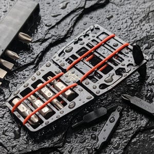 May include: A set of silver-colored screwdriver bits and tools, neatly organized in a dark gray metal case. The bits are secured with orange rubber bands. Additional loose bits are scattered around the case.