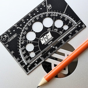 May include: A black rectangular drafting tool with white markings, including a protractor, circular cutouts, and the text MRF DESIGNER metric. An orange pencil with a sharpened lead tip is positioned diagonally across the tool.