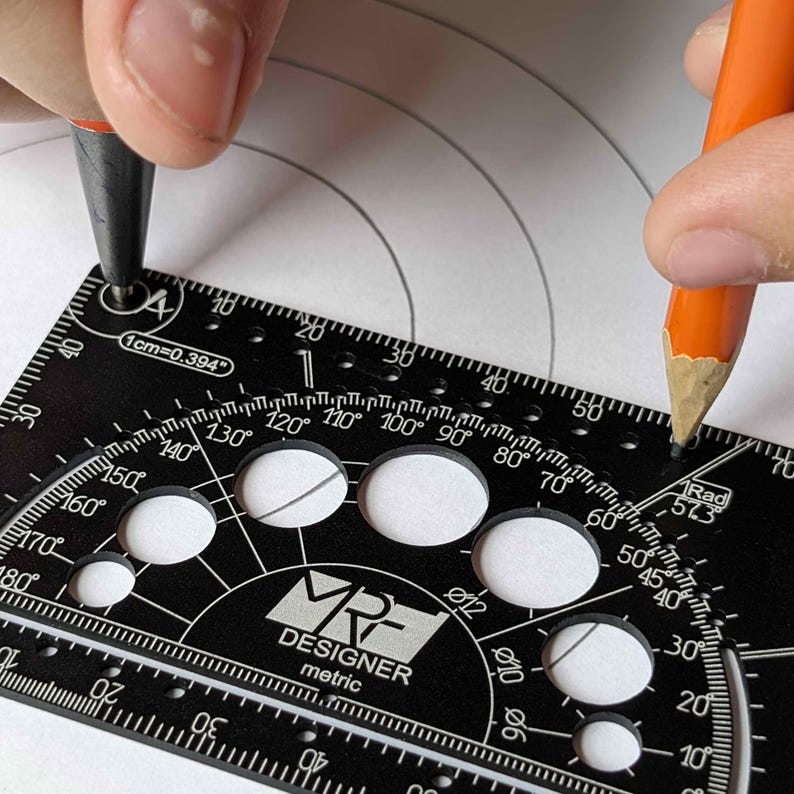 May include: A black MRF Designer metric drafting tool featuring a protractor, circular cutouts, and measurement markings. The tool displays text including 1cm=0.394, 1Rad 57.3, and various degree and millimeter labels, used here with a pencil.