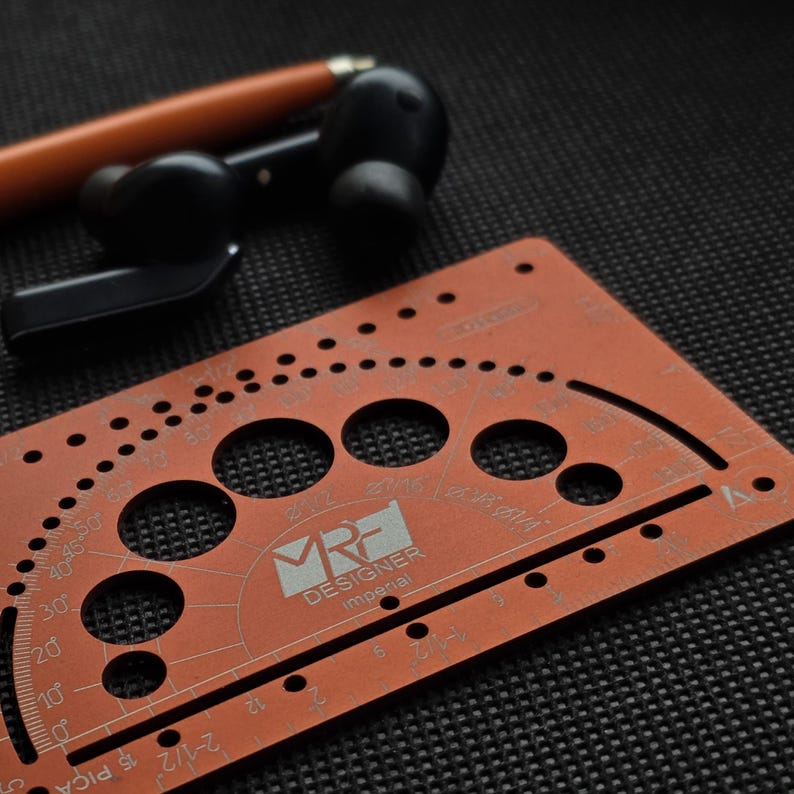 May include: A copper-colored metal drafting template featuring a protractor, circular cutouts, and imperial measurement markings. The tool is labeled with the text MRF DESIGNER imperial. An orange pen and black wireless earbuds are nearby.