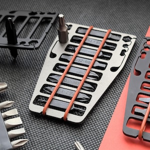 Puede incluir: Un conjunto de accesorios multiherramienta de metal plateado y negro. La herramienta principal tiene múltiples ranuras que contienen varias puntas de destornillador, aseguradas por bandas elásticas naranjas. Se ven herramientas de metal negro adicionales, junto con puntas sueltas.