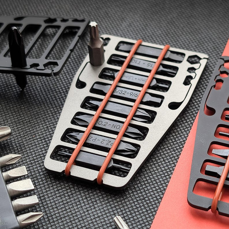 May include: A silver and black multi-tool with various screwdriver bits. The bits are held in place with orange rubber bands. Additional black metal tools are visible in the background. The tools are on a textured surface.