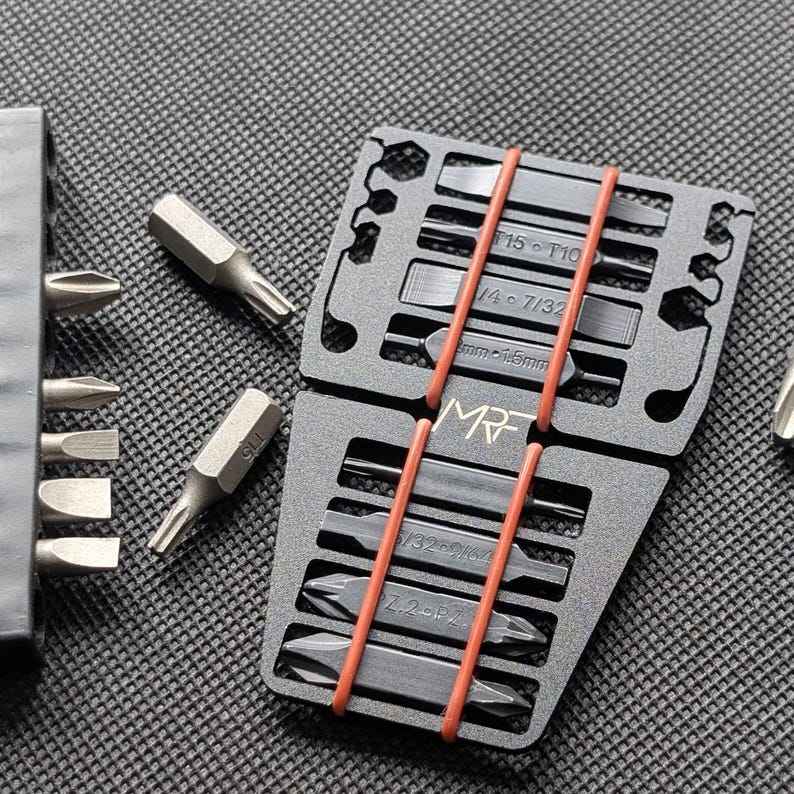 May include: A set of silver and black screwdriver bits, organized in a black holder and secured with orange rubber bands. The holder has labels with size specifications. The brand name "MR" is visible.