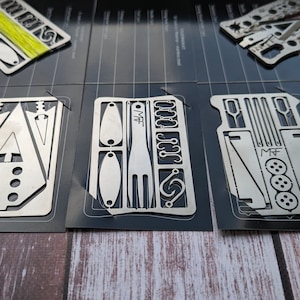 Può includere: Primo piano di tre multitool in metallo con vari strumenti e gadget, tra cui una forchetta, ganci e un righello. Gli strumenti sono realizzati in acciaio inossidabile e hanno una finitura spazzolata.