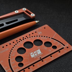 May include: An orange metal drafting tool featuring a protractor, circular cutouts, and a ruler with the text MRF DESIGNER metric. An orange pen and a black and orange tool with screws are positioned nearby on a textured black surface.