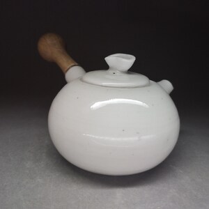 Może przedstawiać: Biały ceramiczny czajniczek z gładkim, błyszczącym wykończeniem i zaokrąglonym korpusem. Posiada krótki, prosty drewniany uchwyt wystający z boku oraz pasującą białą pokrywkę z małym, wyprofilowanym uchwytem w kształcie gałki i małą wylewkę.