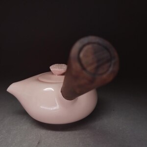 Op de afbeelding: Een lichtroze keramische theepot met een glad, rond lichaam en een korte, donkerbruine houten zijhandgreep. De theepot heeft een bijpassend roze deksel met een kleine, getextureerde, bloemvormige knop in het midden en een korte, puntige tuit.