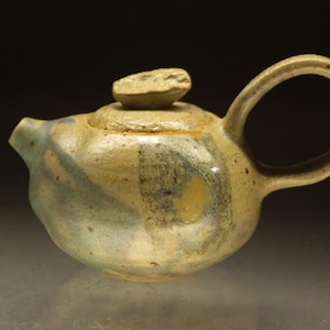 Może przedstawiać: Ręcznie robiony ceramiczny czajniczek z pokrywką i uchwytem. Czajniczek jest głównie beżowy z niebieskimi i żółtymi akcentami. Uchwyt to duża pętla, a pokrywka ma mały, teksturowany guzik. Czajniczek jest prawdopodobnie używany do parzenia herbaty.