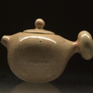 Può includere: Piccola teiera in ceramica rosa chiaro, con corpo arrotondato e coperchio. Ha un beccuccio, un manico e un piccolo pomello. La teiera è probabilmente fatta a mano, con una forma organica unica.