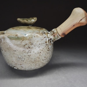 Op de afbeelding: Een keramische theepot met een gespikkeld, gebroken wit en lichtbruin glazuur, met een onregelmatig, getextureerd oppervlak. De theepot heeft een bijpassend deksel met een kleine knop en een zijhandvat van lichtgekleurd, glad, gepolijst hout.