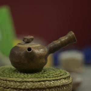 Può includere: Una teiera in ceramica marrone con un corpo rotondo e un beccuccio. La teiera ha un manico e un coperchio con un piccolo pomo. La teiera è appoggiata su un tappetino intrecciato.