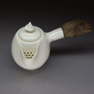 Può includere: Una teiera in ceramica bianca con manico in legno. La teiera ha un corpo arrotondato, un beccuccio con piccoli fori e un coperchio con un piccolo pomello. Il manico è in legno dall'aspetto naturale e strutturato.