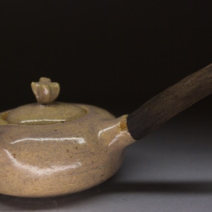 May include: A handmade ceramic teapot with a lid and a long, dark wooden handle. The teapot is a muted beige color with a small spout and a flower-shaped knob on the lid. The handle is dark brown.