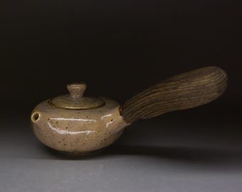 May include: A small, round, ceramic teapot with a long, wooden handle. The teapot is a speckled beige color with a matching lid. The handle is a light brown color. The teapot has a small spout.