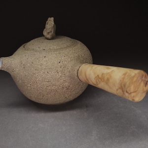 Op de afbeelding: Een ronde, beige keramische theepot met een korrelig, gespikkeld oppervlak, een kleine sculpturale knop op het deksel en een gladde, lichtgekleurde houten handgreep aan de zijkant. De tuit heeft een witte afwerking aan de binnenzijde.