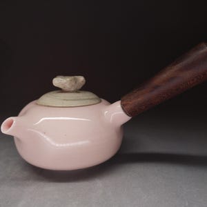 Może przedstawiać: Jasnoróżowy ceramiczny czajniczek o gładkim, błyszczącym wykończeniu. Czajniczek posiada boczny uchwyt z ciemnobrązowego drewna z widocznym usłojeniem, beżową pokrywkę o teksturowanej powierzchni oraz mały, rzeźbiony uchwyt na szczycie pokrywki.