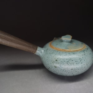 Może przedstawiać: Ceramiczny czajniczek z nakrapianą jasnoniebieską glazurą i gładką, ciemnobrązową drewnianą rączką boczną. Czajniczek ma okrągły korpus i pasującą pokrywkę z małym uchwytem, prezentując rustykalną fakturę oraz naturalne, ziemiste odcienie kolorów.