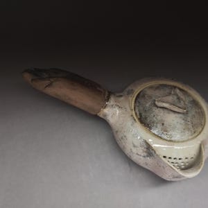 Może przedstawiać: Ceramiczny czajniczek z jasnym, nakrapianym szkliwem i brązowym uchwytem w kształcie ludzkiej dłoni. Czajniczek posiada okrągłą pokrywkę z małym trójkątnym uchwytem oraz wbudowane sitko przy dzióbku, wykazując teksturowane wykończenie powierzchni.