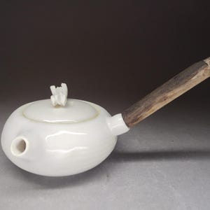 Może przedstawiać: Biały ceramiczny czajniczek o gładkim, zaokrąglonym korpusie i krótkim dzióbku. Pokrywka posiada mały, rzeźbiarski biały uchwyt. Długa, prosta, teksturowana drewniana rączka wystaje z boku czajniczka, tworząc kontrast z ceramiczną powierzchnią.