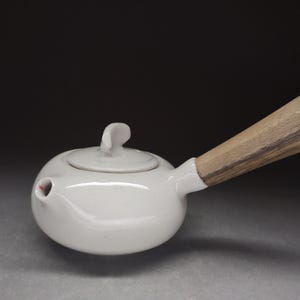 Op de afbeelding: Een witte keramische theepot met een gladde, glanzende afwerking, voorzien van een rond lichaam, een kleine tuit, een bijpassend deksel met een gebogen handvat en een lange, cilindrische houten zijgreep met zichtbare houtnerf.