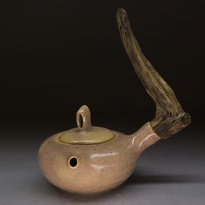 May include: A handcrafted, ceramic teapot with a light brown glaze. The teapot features a natural wood handle and a small, round lid. The design includes a spout and a small hole on the side. The overall aesthetic is rustic and organic.