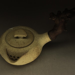 Op de afbeelding: Een beige keramische theepot met een gespikkelde textuur, een rond deksel met een kleine knop en een donkerbruin, knoestig houten takhandvat. De theepot is voorzien van een ingebouwde zeef met meerdere ronde gaten bij de tuit.