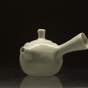 Può includere: Una piccola teiera in ceramica bianco sporco con corpo arrotondato, beccuccio e manico. La teiera ha un coperchio con un piccolo pomello. Lo sfondo è nero e la teiera è appoggiata su una superficie riflettente.