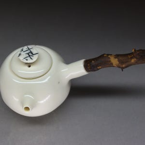 Könnte beinhalten: Eine weiße Keramik-Teekanne mit einem einzigartigen Design. Sie hat einen runden Körper, einen kleinen Ausguss und einen Deckel mit schwarzen Markierungen. Der Griff ist ein natürlich aussehender, dunkelbrauner Ast.