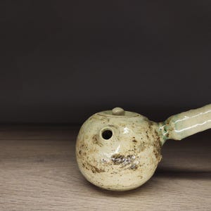Può includere: Una teiera in ceramica rotonda color bianco sporco con manico laterale e un piccolo coperchio. La superficie presenta una consistenza maculata e screziata con toni terrosi marrone scuro e verde, conferendole un aspetto organico e vissuto.