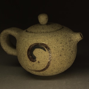 Peut inclure: Une théière ronde en céramique beige mouchetée avec une surface texturée et granuleuse. Elle comprend une petite anse, un bec court et un couvercle assorti avec un bouton. Un motif en spirale peint à la main en brun foncé est centré sur le corps.