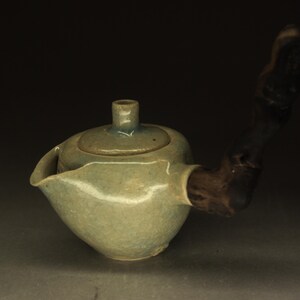 Può includere: Una teiera in ceramica con corpo smaltato verde salvia chiaro e un manico in legno scuro e testurizzato che si estende lateralmente. La teiera presenta un coperchio arrotondato con un piccolo pomello centrale e un beccuccio largo e aperto.