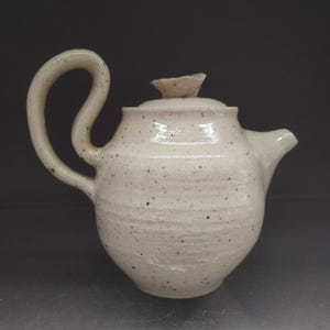 Può includere: Una teiera in ceramica bianca con una glassa macchiettata e un'unica maniglia curva. La teiera ha un piccolo beccuccio e un coperchio con un pomo.
