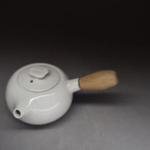 Op de afbeelding: Een witte keramische theepot met een glad, rond lichaam, een kleine tuit en een deksel met een textuurrijke, bloemvormige knop. De theepot is voorzien van een lichtgekleurd, cilindervormig houten handvat aan de zijkant van de pot.